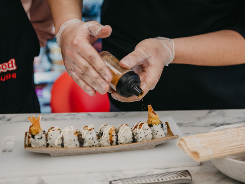 Close up of hands in food prep gloves sprinkling sushi seasoning onto a rectangular dish of California rolls 